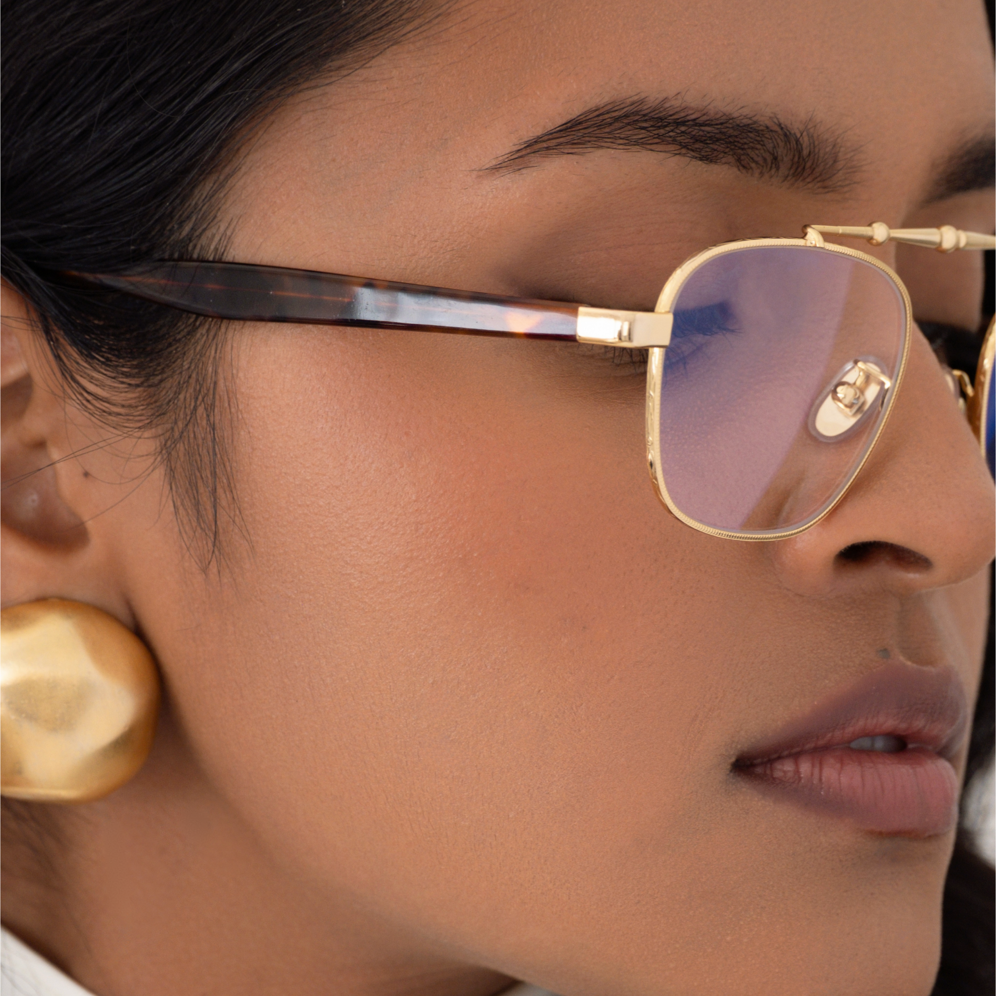 Close-up of a person wearing the balaustra aviator blue light glasses. Details shown include the tortoise acetate temple arm on the sides, and the gold frame from the front which has intricate gold details around the lenses and a sculptural top bar, inspired by architectural railings.