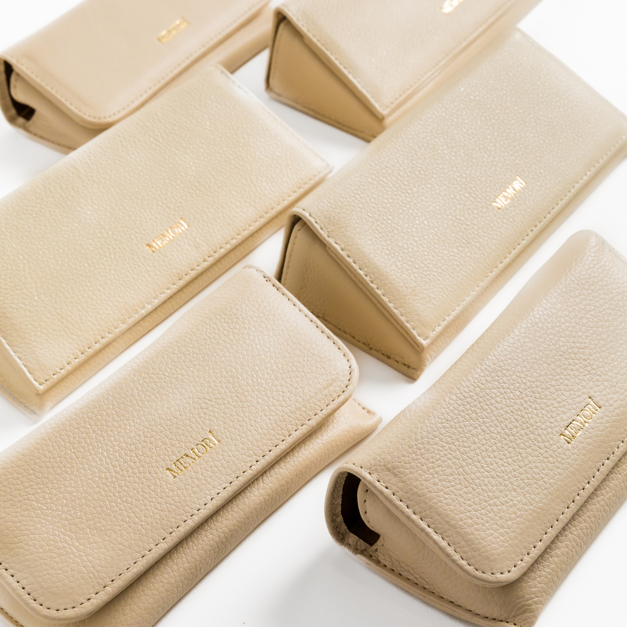 Photo depicting 6 different Memori brand sunglass cases arranged on a table. two different sizes, but all cases are made of the same cream/ivory pebbled leather and feature a gold embossed foil logo in the lower center of the folding flap. The cases appear to be very luxurious, protective, and great quality.