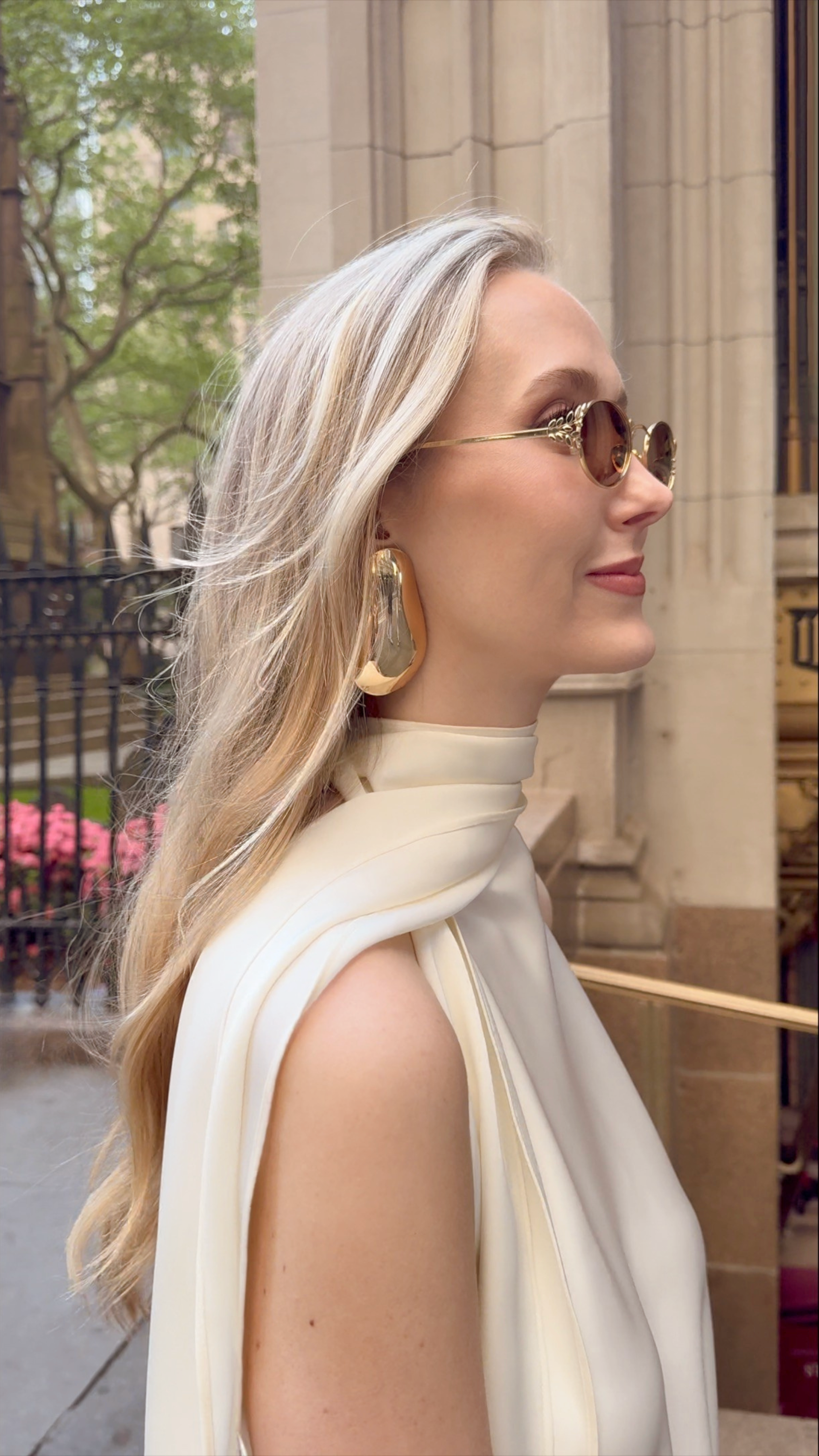 Ashlyn Lawrence, Founder of Centre Piece, wearing a top of her own design on the streets of NYC paired with oversized gold earrings and Memori Laurel oval sunglasses in gold with warm brown lenses. The overall aesthetic is very elegant and warm, and the metal leaf side details of the glasses look very elegant. 