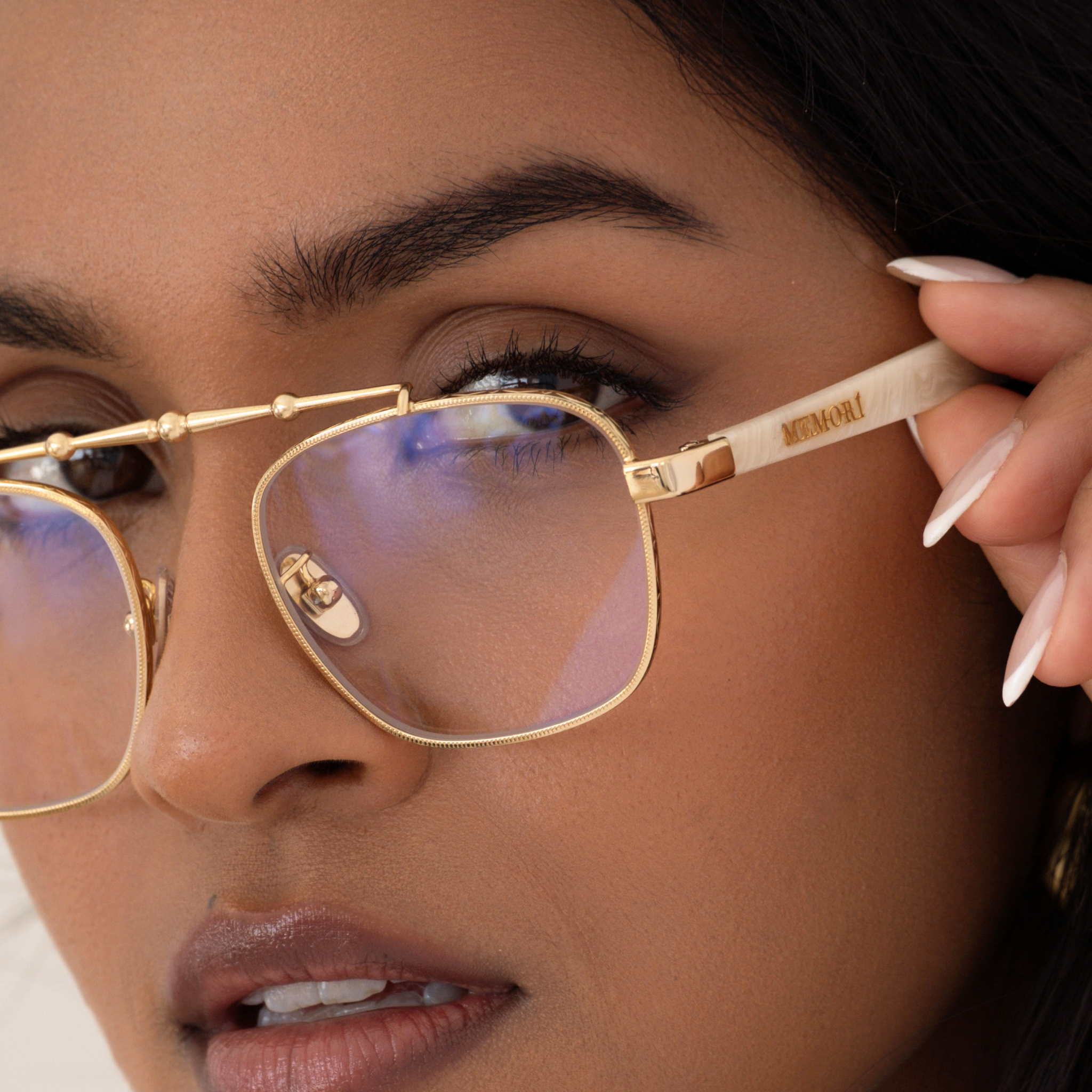 Close-up of model wearing Memori balaustra aviator glasses, showing the subtle texture around the gold rim of the lens, the metal brow bar, and the Memorí logo in gold on the external side temple. 