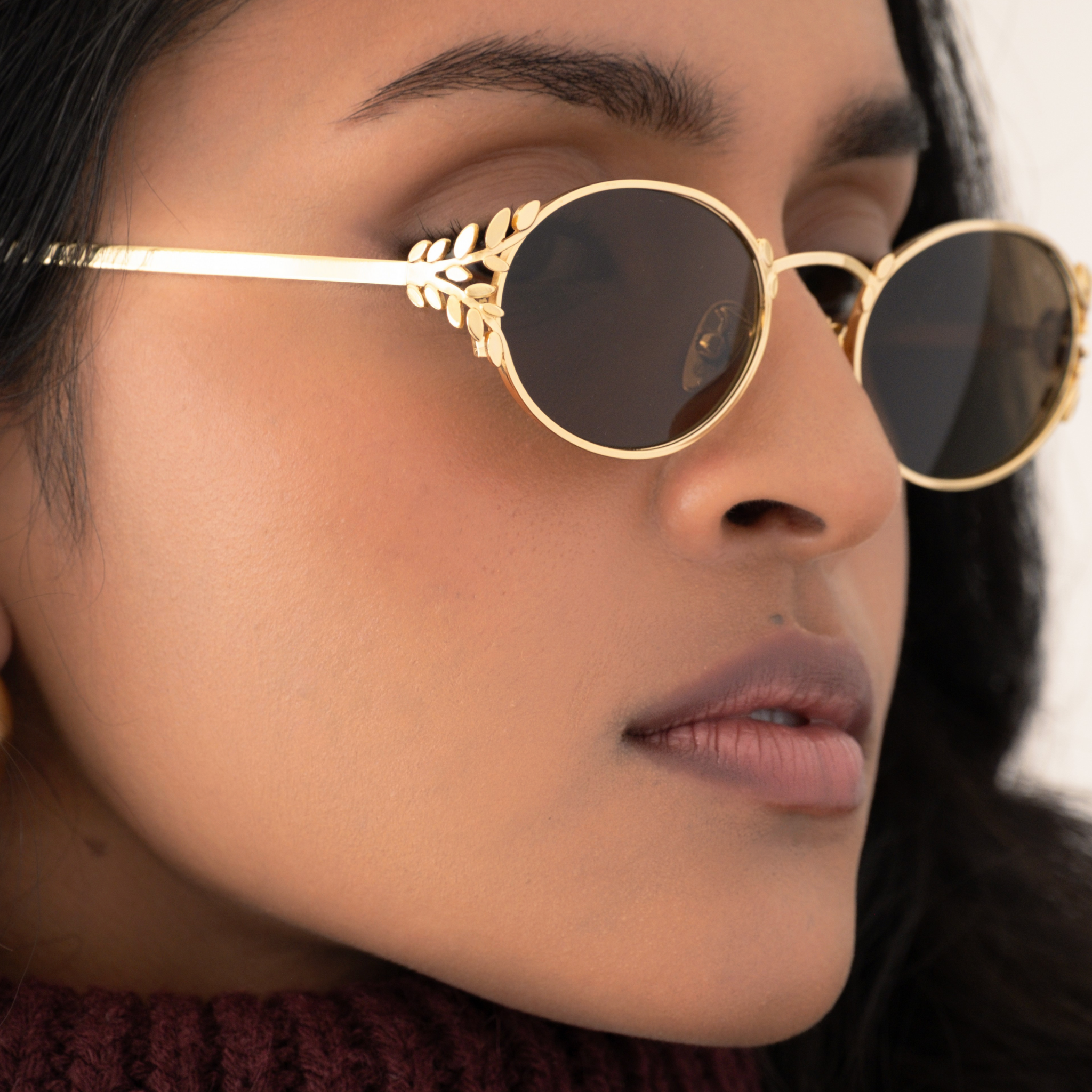 Close-up of a person with a small face shape wearing memori Laurel oval sunglasses in gold. Photo focuses on the detailed laurel leaf sides, the glasses look like a combination of sunglasses and fine jewelry for the face.