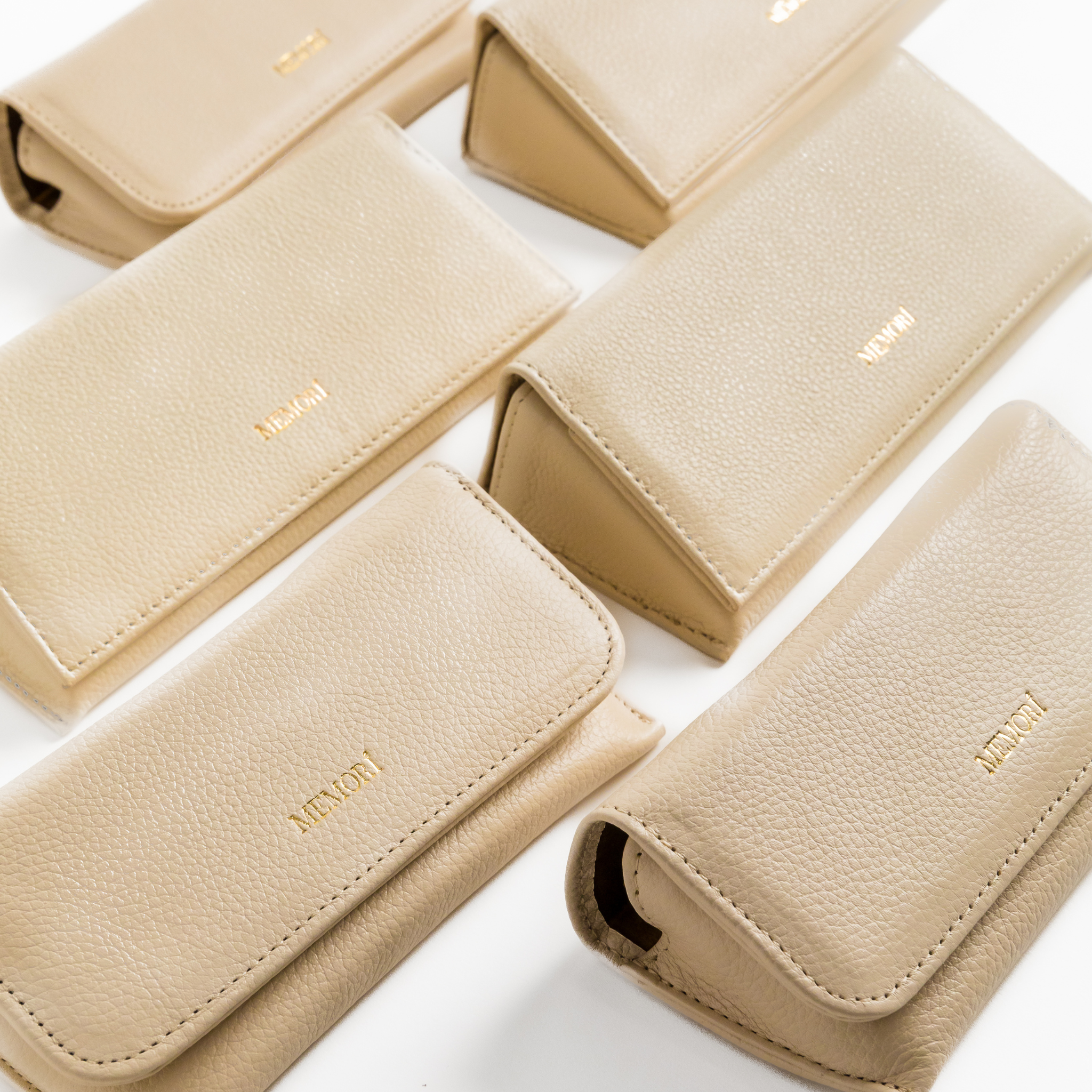 Photo depicting 6 different Memori brand sunglass cases arranged on a table. two different sizes, but all cases are made of the same cream/ivory pebbled leather and feature a gold embossed foil logo in the lower center of the folding flap. The cases appear to be very luxurious, protective, and great quality. 