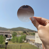 A hand holding up an uncut sunglass lens - brown gradient color with sunlight from Italy streaming through the lens. 