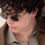 Male model wearing Laurel Oval sunglasses in silver and a brown leather jacket, looking down. This photo offers a macro view of the laurel leaf side details. the leaves are separated and shining in the light. 