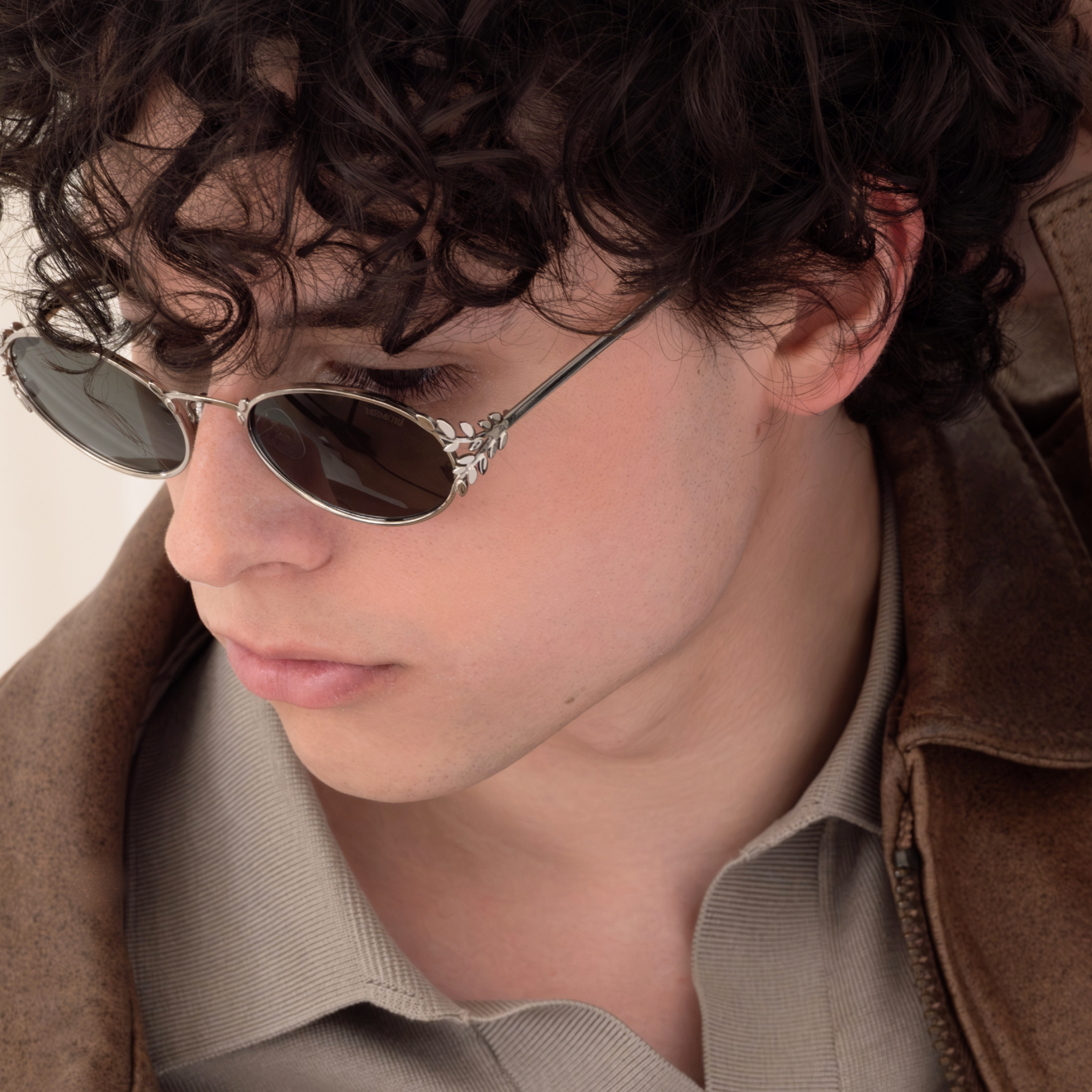 Male model wearing Laurel Oval sunglasses in silver and a brown leather jacket, looking down. This photo offers a macro view of the laurel leaf side details. the leaves are separated and shining in the light. 