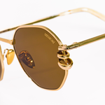 Zoomed in view of metal details of these gold pierced hexagonal sunglasses. Rim has a leaf detail pressed into the metal, "Memorí" logo is etched in the upper corner of the lens, and two chunky hoop piercings are made through the lens of the sunglasses. 